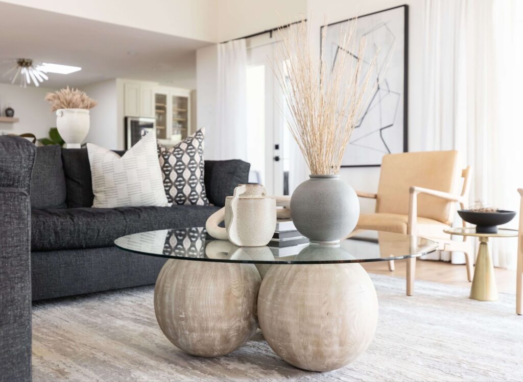 A modern living room featuring a comfortable black sofa and an elegant glass coffee table adorned with vases. Wooden spheres are scattered on a patterned rug in front of the sofa, and a woman sits on the sofa, enjoying the luxury of the living room.
