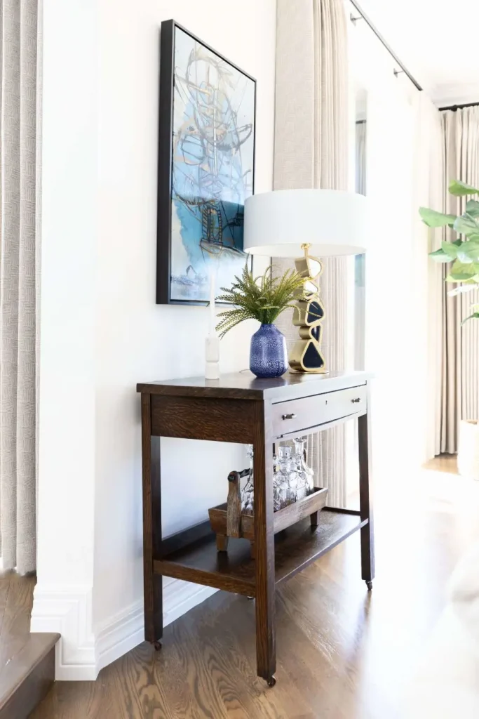 Dark wood console table with a white lamp, blue vase with green plants, and abstract wall art