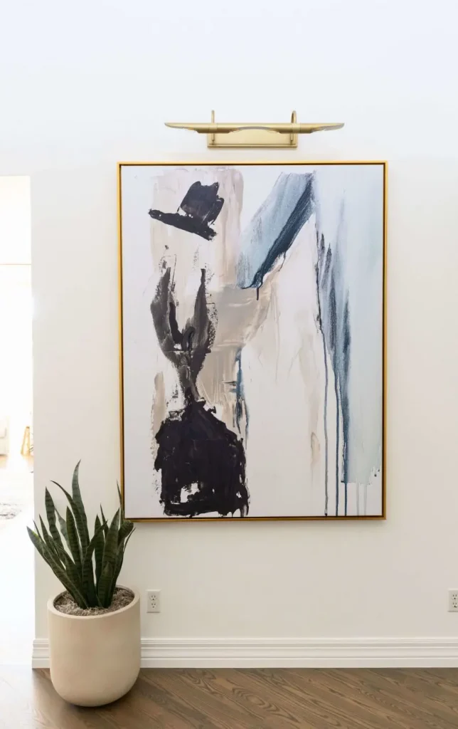 Abstract painting with black, beige, and blue brushstrokes in a gold frame, mounted on a white wall above a potted snake plant