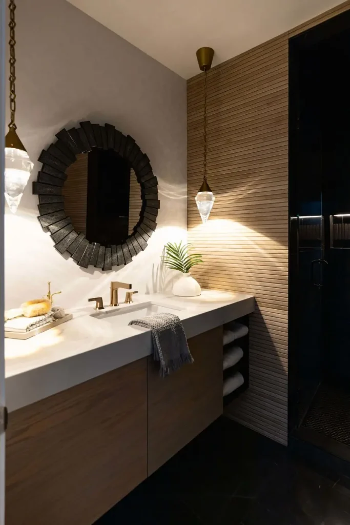 Bathroom vanity with wooden cabinets, white countertop, gold faucet, round black mirror, hanging pendant lights, and a small plant in a white vase.