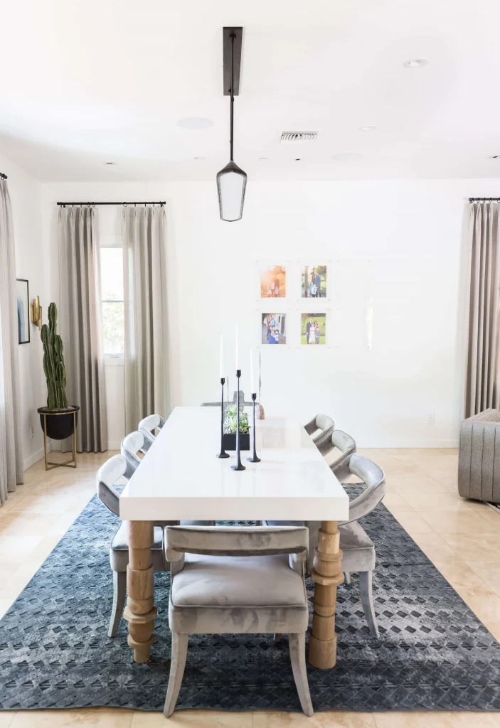 Bright dining room with a long white table, six gray velvet chairs, black candle holders, and beige curtains.