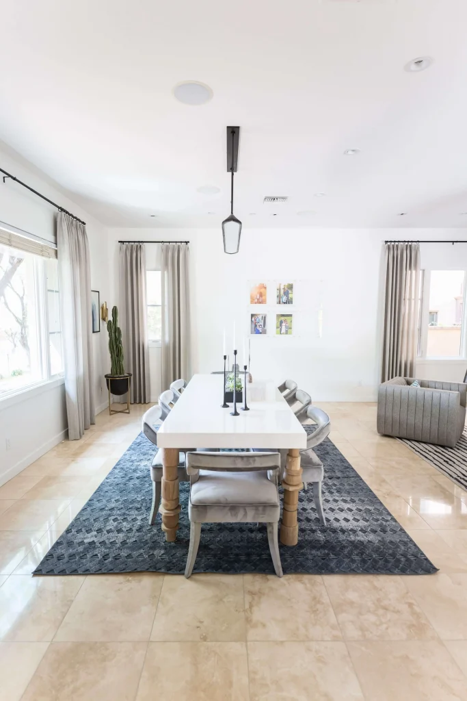 Bright dining room with long white table, gray velvet chairs, beige curtains, and cactus plant.