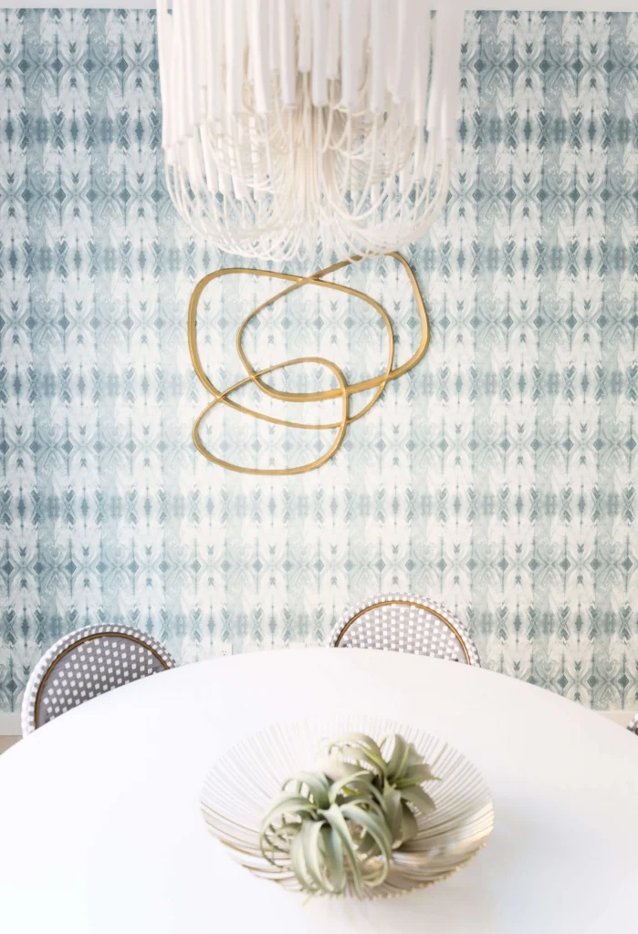 Abstract gold metal wall art hanging on a blue patterned wallpaper behind a white round table with plants in a decorative bowl.