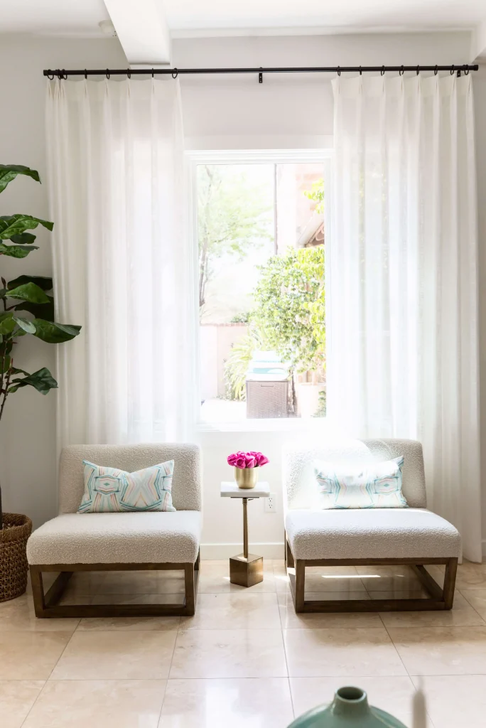 Two modern white armchairs with patterned cushions, small gold side table with pink flowers, and sheer white curtains
