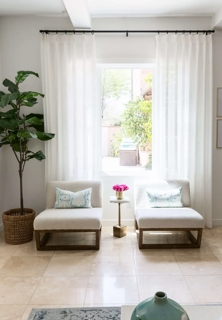 Two modern white armchairs with patterned cushions, small gold side table with pink flowers, sheer white curtains, and a potted plant on the left.