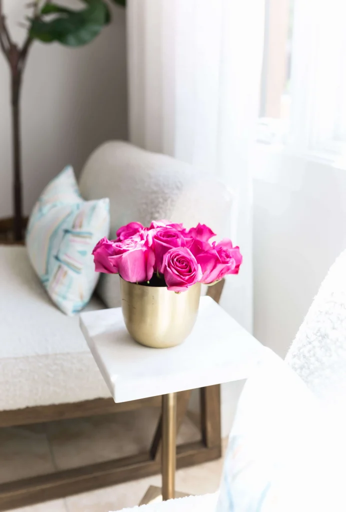 Close-up of a gold vase with bright pink roses on a small marble-topped side table next to a white armchair with a patterned cushion.