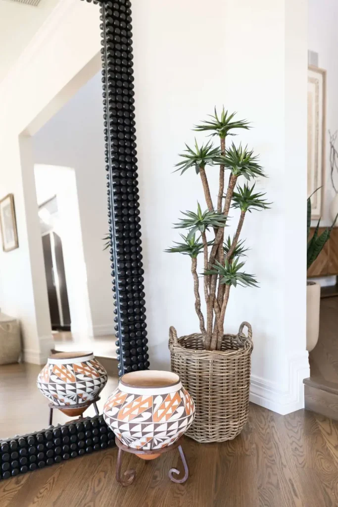 Colorful pot on stand beside a potted tall plant in woven basket with black round-studded mirror behind