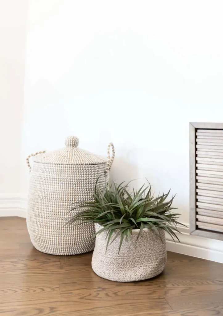 Large woven basket with lid and handles next to a smaller woven basket holding a green leafy plant on wooden floor