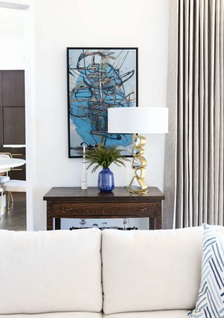 Wooden console table with abstract blue art above and modern gold table lamp beside a blue vase with greenery.