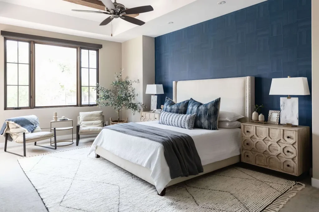 Bright bedroom with blue accent wall, beige upholstered bed, wooden nightstands, and modern seating area