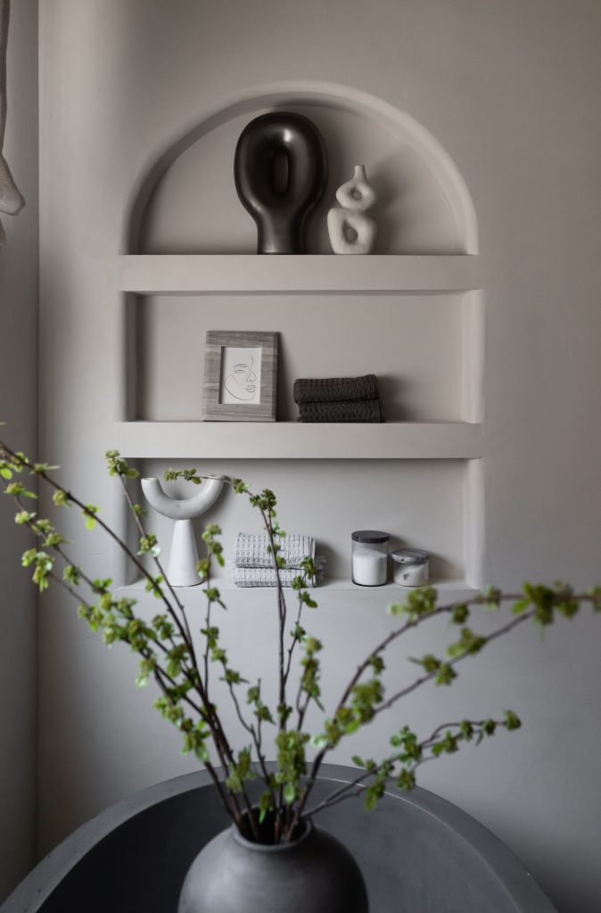 Minimalist wall shelves with abstract sculptures, monochrome decor, towels, candles, and a framed line drawing. A vase of green branches is on a round table in the foreground— Living with Lolo.