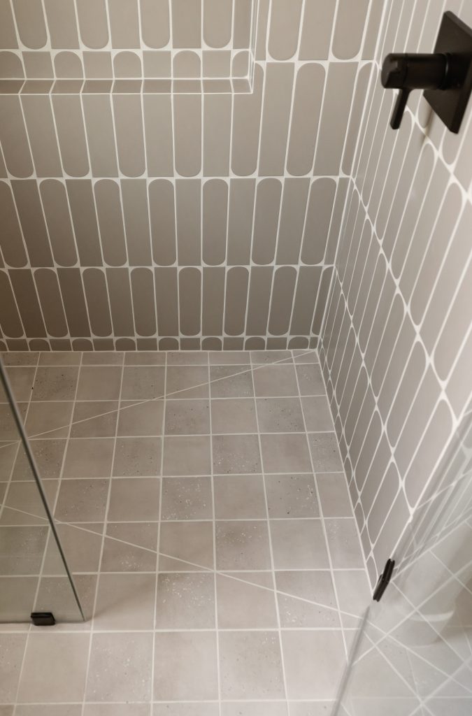 Bathroom with light-colored rectangular wall tiles, square floor tiles, a glass shower partition, and a black metal door hinge—Living with Lolo.