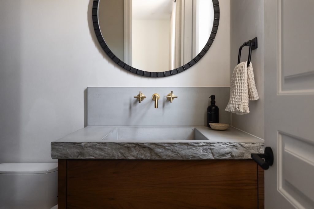 Modern bathroom with a stone countertop, gold faucet, round mirror, black towel holder, and a soap dispenser on the right—Living with Lolo.