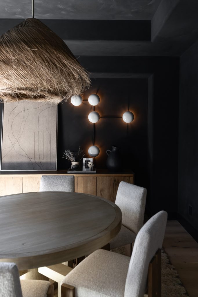 A round wooden dining table with four upholstered chairs is in a dark room with a rattan pendant lamp, abstract art, and a modern wall sconce with four glowing bulbs. — Living with Lolo.
