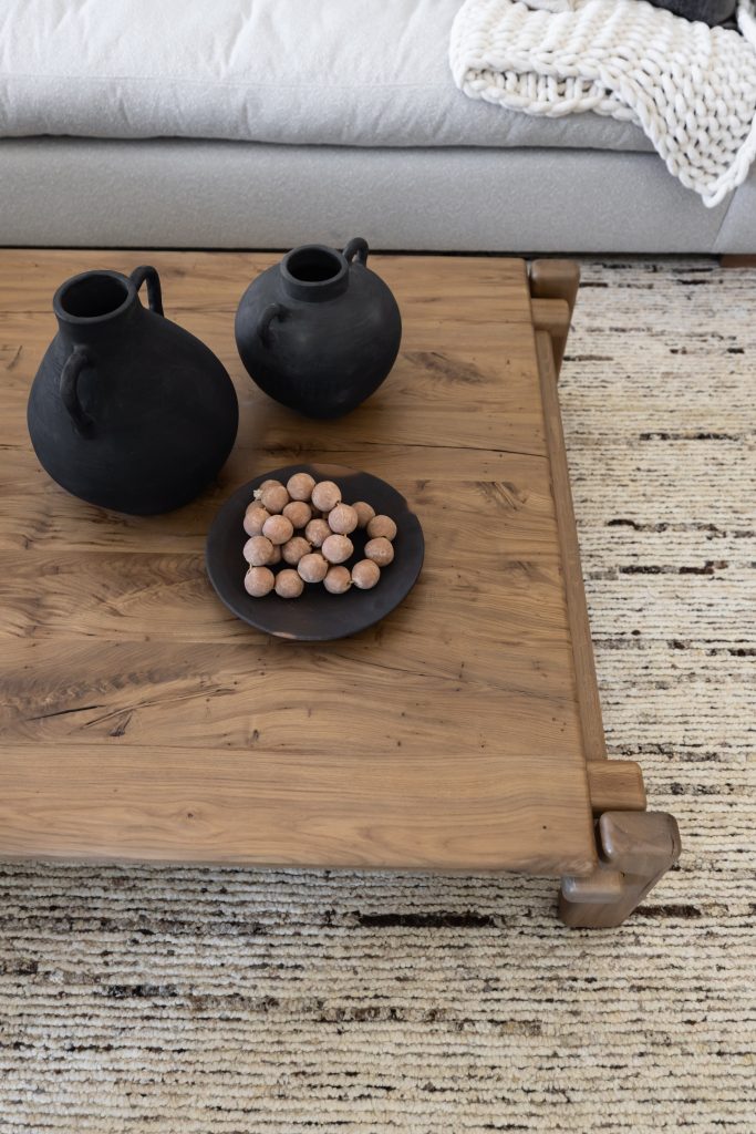 A wooden coffee table sits on a cream and brown textured rug, with two black ceramic pitchers and a plate of snacks. — Living with Lolo.