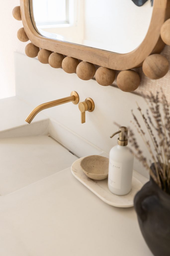 A bathroom sink features a gold wall-mounted faucet, wooden-framed mirror, soap dispenser, dish, and vase with dried lavender—Living with Lolo.