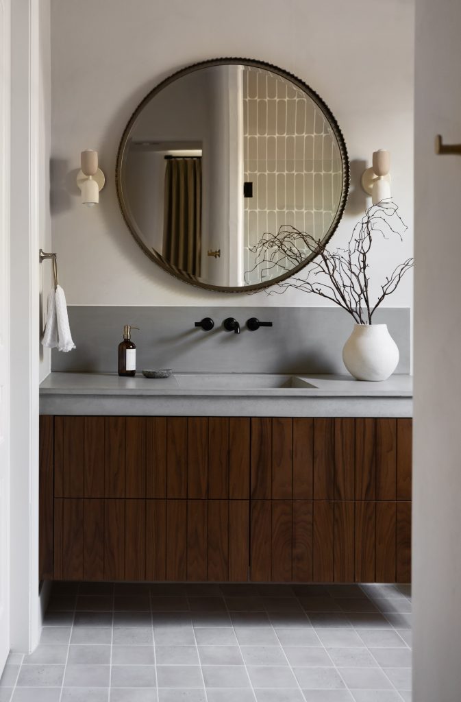 A bathroom with a round mirror, wall-mounted faucets, wood vanity, gray countertop, white vase with branches, and tile flooring.— Living with Lolo.