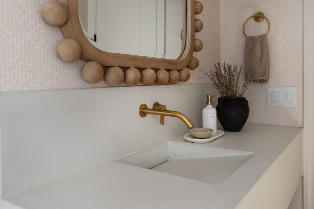 A bathroom sink with a gold wall-mounted faucet, a mirror, black vase with dried plants, soap dispenser, and beige hand towel. — Living with Lolo.