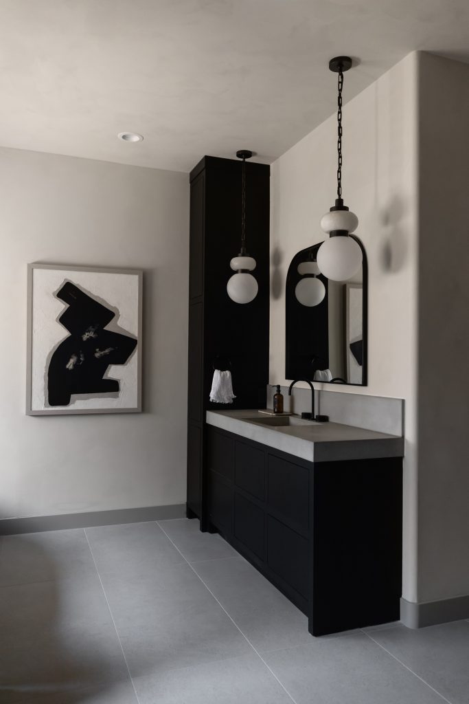 Modern bathroom with black vanity, rectangular mirror, globe pendant lights, abstract artwork, and light gray tile flooring—Living with Lolo.