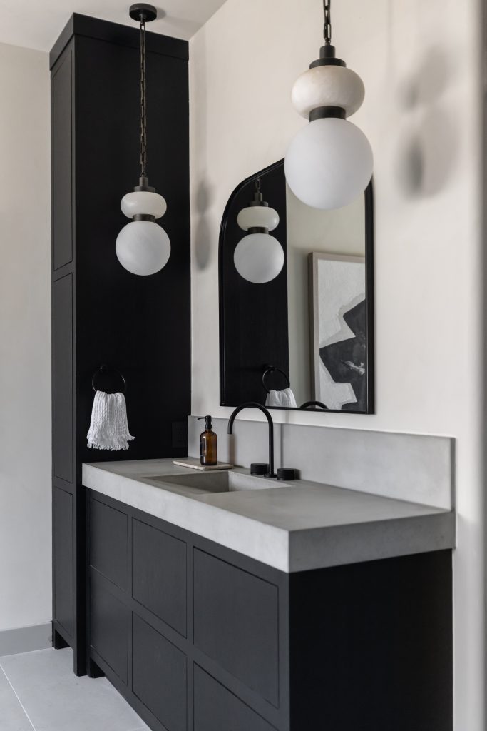 Modern bathroom with a black vanity, concrete countertop, round mirror, pendant lights, tassel wall sconce, and soap dispenser.— Living with Lolo.