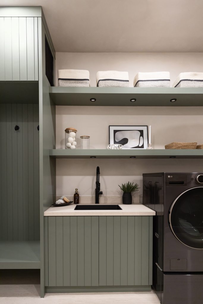Modern laundry room with sage green cabinets, open shelves with baskets, a black faucet, small sink, and a front-load washer—Living with Lolo.