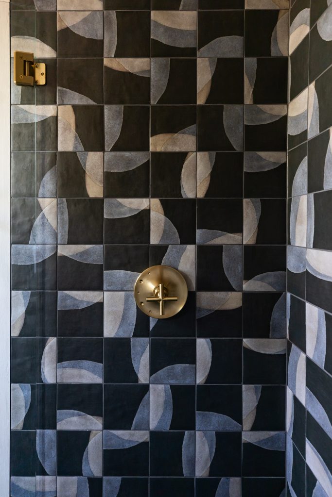 Shower wall with dark geometric ceramic tiles featuring curved light gray patterns and a central brass fixture with a round handle.— Living with Lolo.