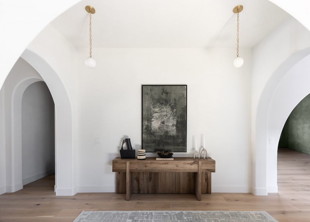 A wooden console table with decor and framed artwork sits against a white wall between two arched doorways on light wood floors—Living with Lolo.