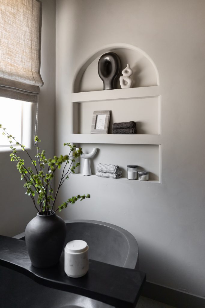 Minimalist bathroom with built-in arched shelves, a dark counter holding a vase with green branches, and a nearby bathtub— Living with Lolo.