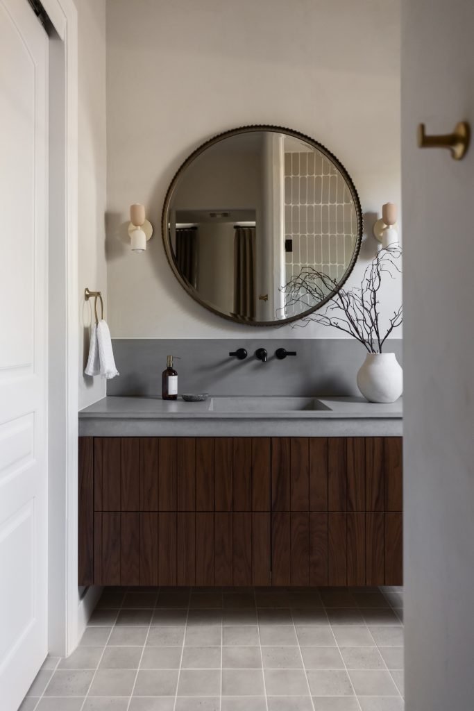 A round mirror hangs above a wall-mounted faucet and wooden vanity with a gray countertop; potted branches sit on top. Neutral tile flooring completes the space. — Living with Lolo.