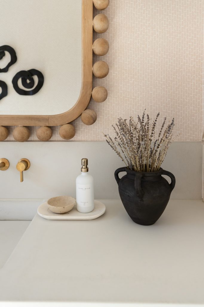A black vase with dried lavender, a soap dish, and a bottle rest on a white countertop below a rounded wooden mirror.— Living with Lolo.