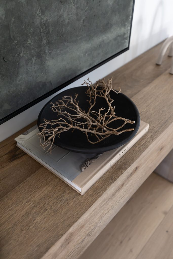 A black bowl with dry twigs is placed on a closed book atop a wooden surface next to a wall—Living with Lolo.
