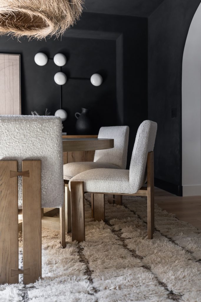 A light wood table and textured cream chairs sit on a shaggy rug, with a dark wall and round wall lights behind—Living with Lolo.