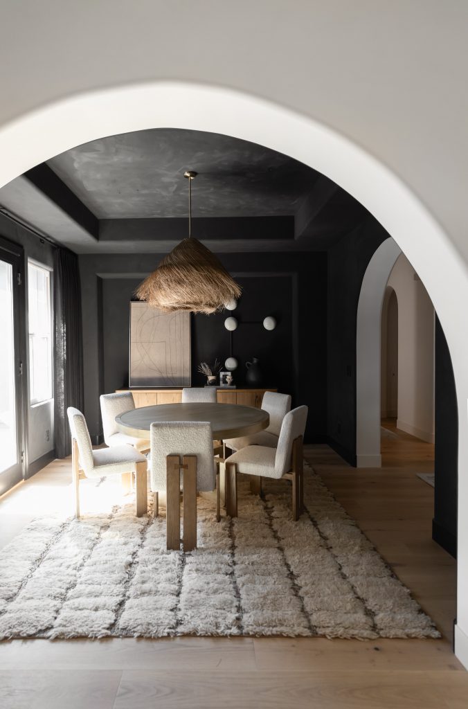 A modern dining room with a round table, four upholstered chairs, a shag rug, woven pendant light, and black accent walls— Living with Lolo.