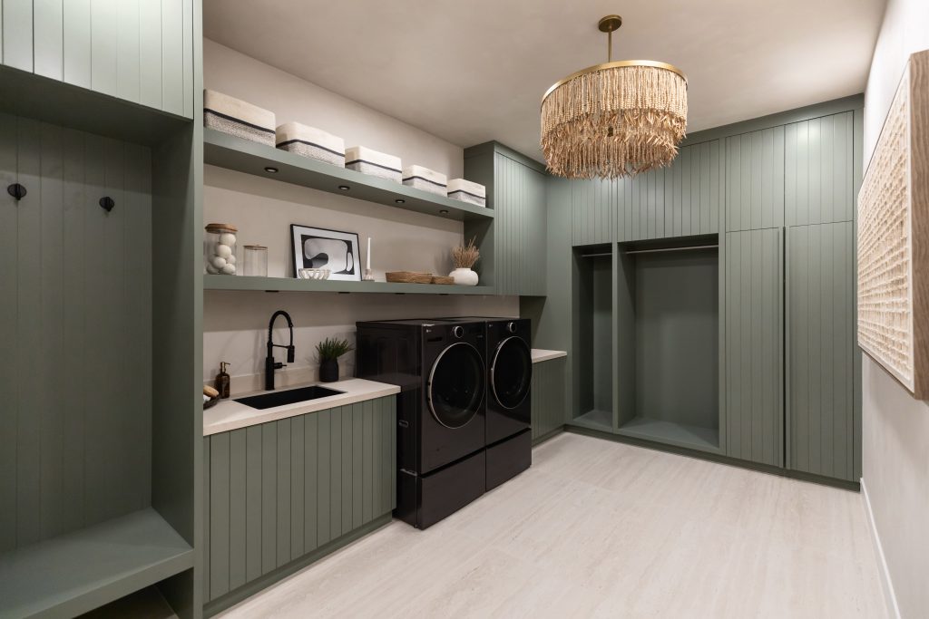 Laundry room featuring green cabinets, black washer and dryer, open shelving with baskets, a sink, and a gold pendant light — Living with Lolo.
