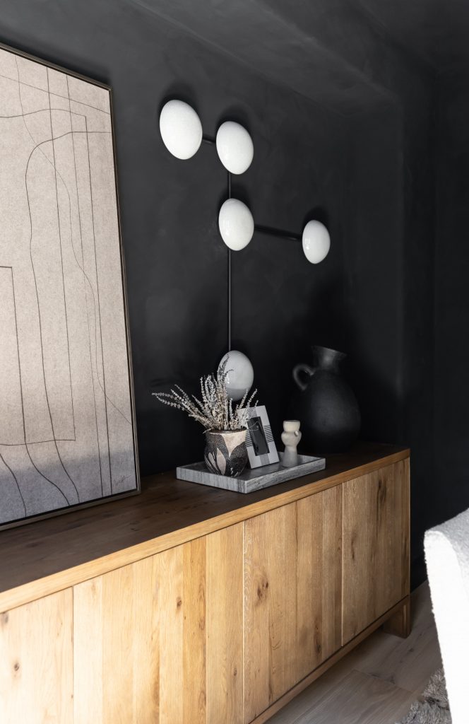 A wooden sideboard stands against a dark wall with a modern lamp, abstract art, and small decorative accents—Living with Lolo.