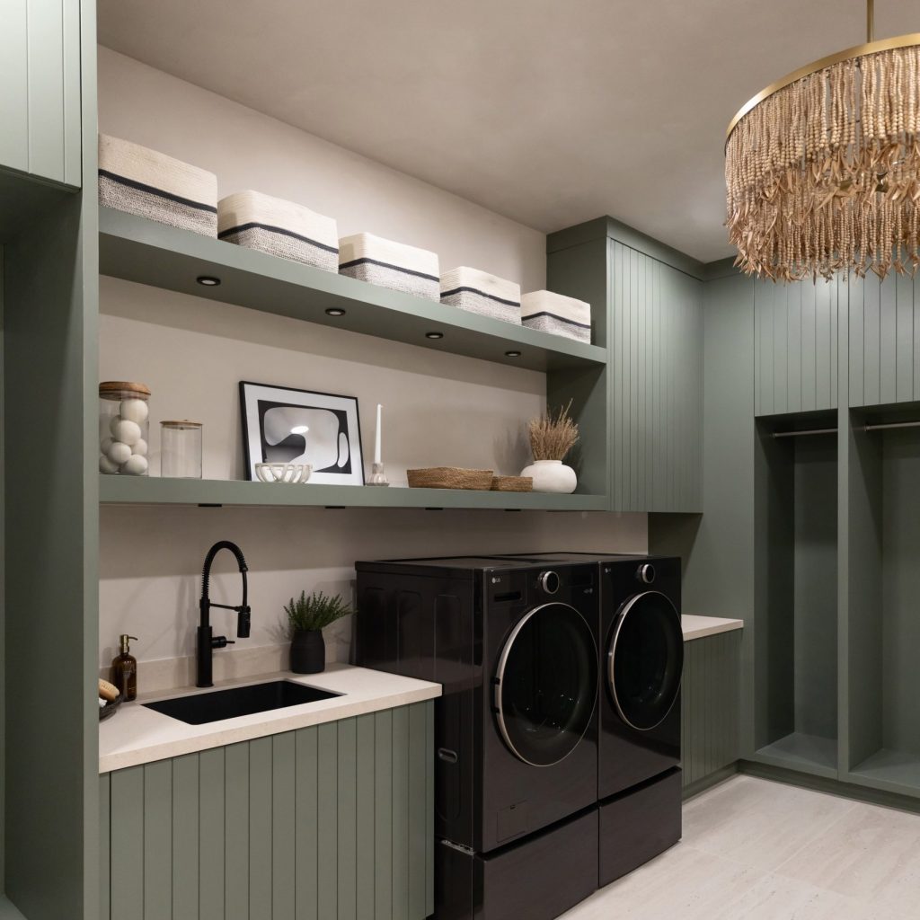 Modern laundry room featuring dark appliances, green cabinets, open shelves with baskets, a small sink, and a fringed chandelier—Living with Lolo.