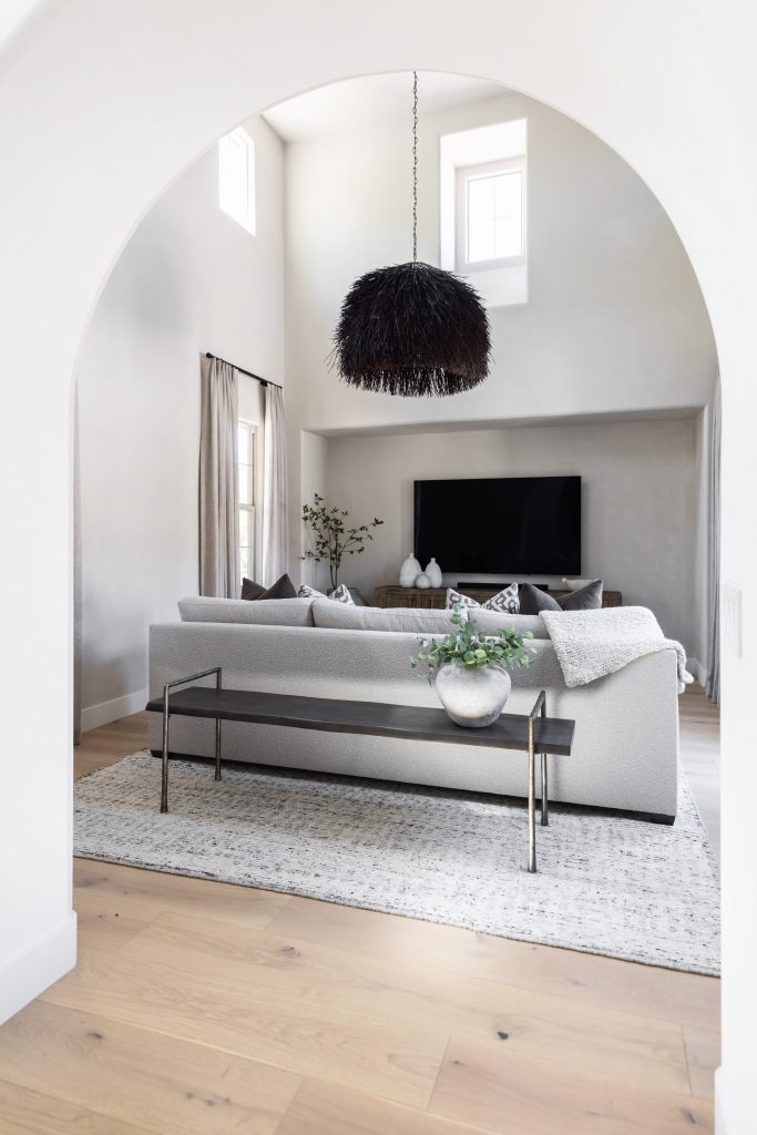 Modern living room with light wood floors, gray sofa, black feathered pendant light, large TV, decorative plant, and arched doorway—Living with Lolo.