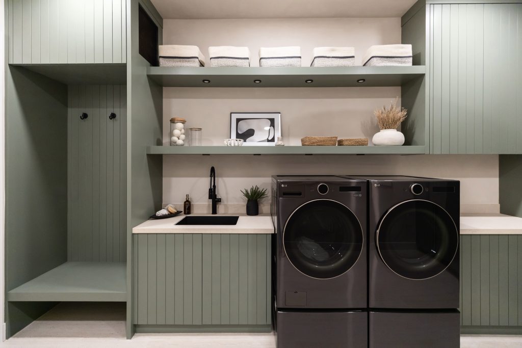 Laundry room with green cabinets, black washer and dryer, a built-in sink, and open shelves with towels and jars— Living with Lolo.