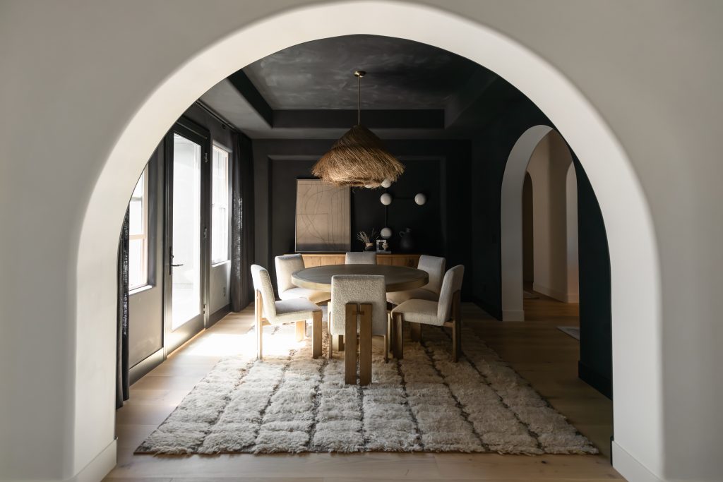 A dining room with a round wooden table, six upholstered chairs, shaggy rug, woven pendant light, and dark walls framed by an archway. — Living with Lolo.