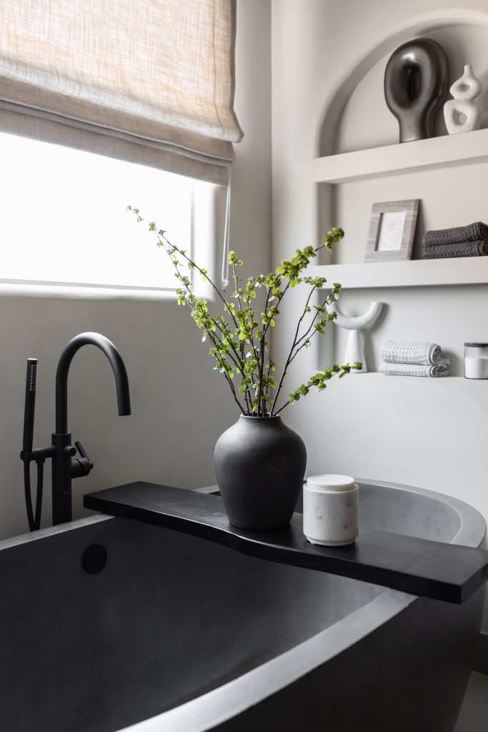 A matte black tub with a black faucet sits near built-in shelves holding a vase with green branches, a candle, and rolled towels — Living with Lolo.