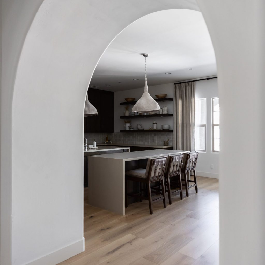 Modern kitchen seen through an arched doorway with an island, barstools, pendant lights, open shelves, and large windows—Living with Lolo.