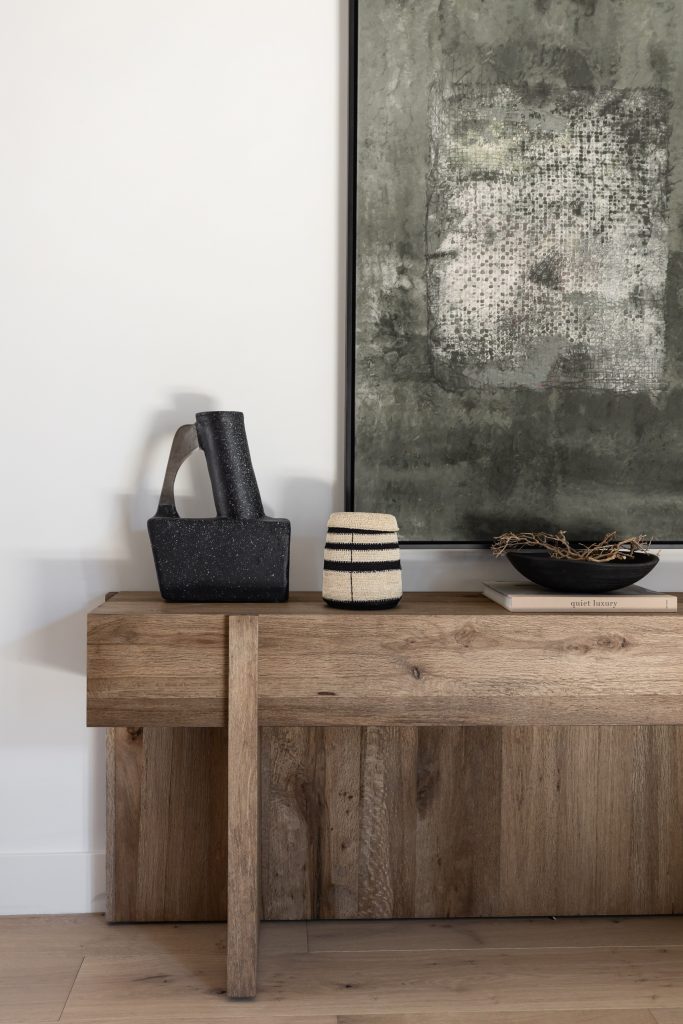 A wooden console table holds black and white decor, a bowl with twigs, and sits below a large abstract painting — Living with Lolo.