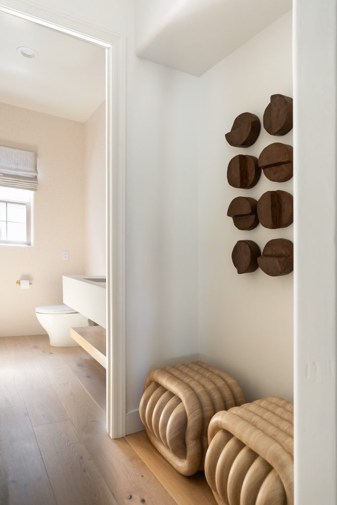 Minimalist hallway with two wooden stools, abstract wood wall art, and a view into a bathroom with light floors and white walls — Living with Lolo.