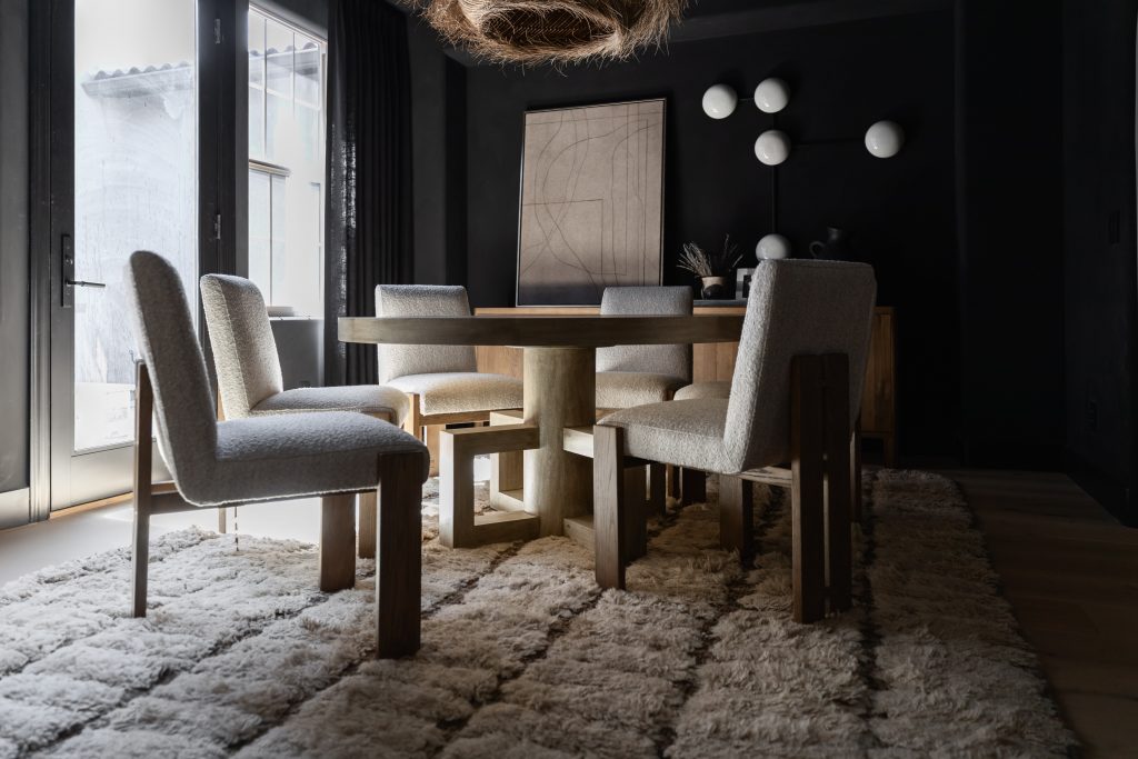 A modern dining room with a round wooden table, six upholstered chairs, a textured rug, abstract art, and globe wall lights. — Living with Lolo.