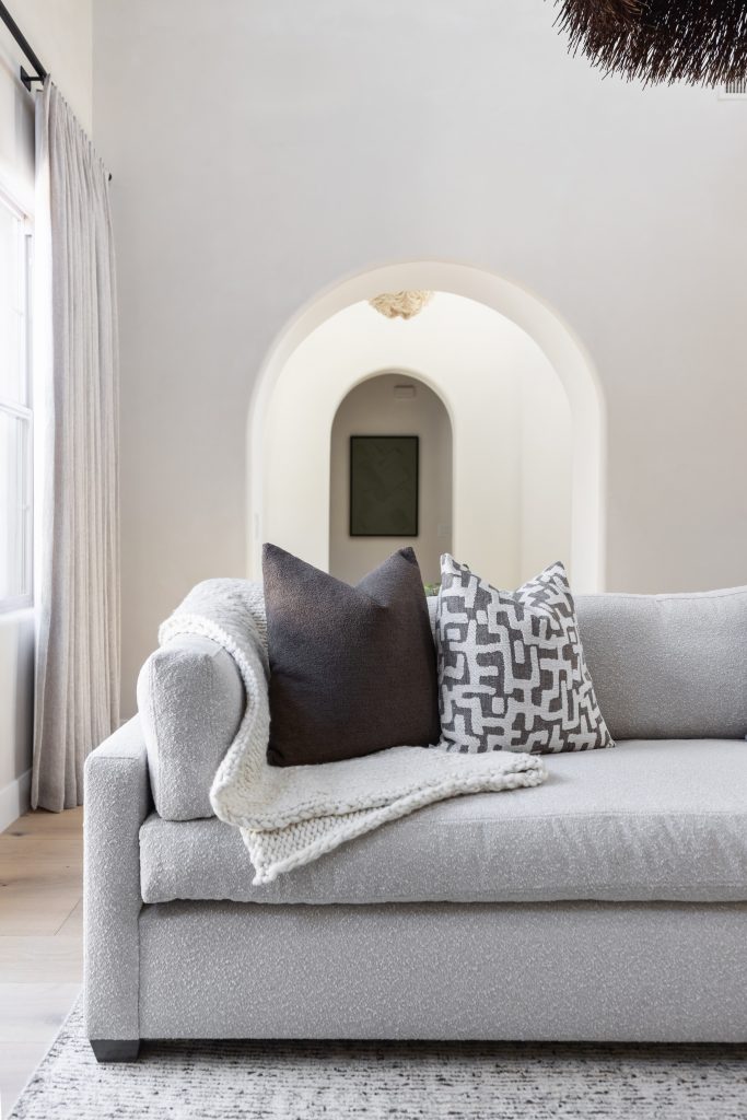 A light gray sofa with two patterned pillows and a knit blanket faces an arched doorway in a bright, minimalist living room—Living with Lolo.