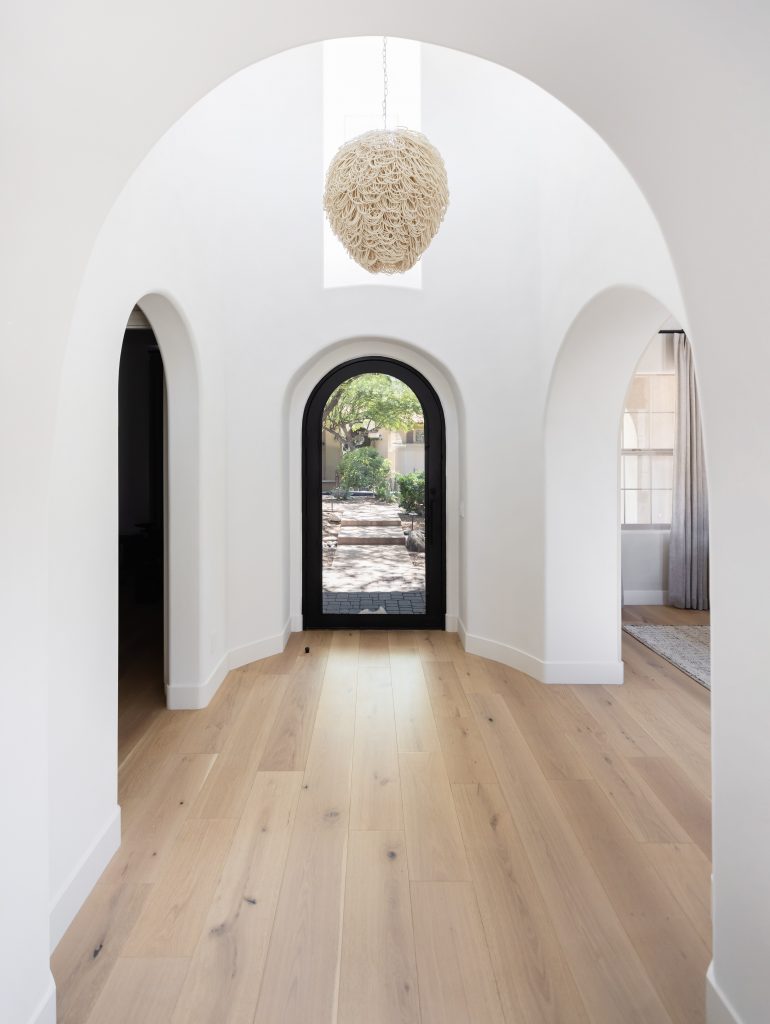 Hallway with light wood floors, white arched walls, a woven pendant light, and a black-framed arched glass door.— Living with Lolo.