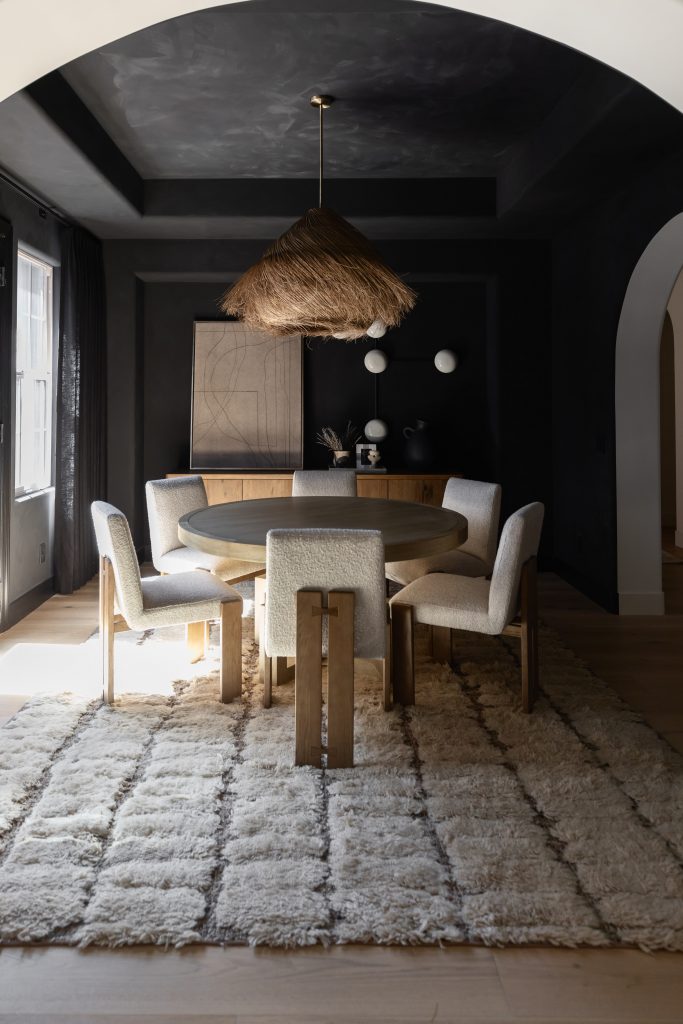 A modern dining room with a round wooden table, six upholstered chairs, a shaggy rug, a woven pendant light, and dark accent walls. — Living with Lolo.
