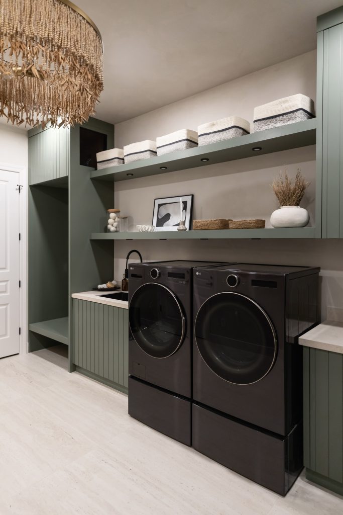Modern laundry room with green cabinets, open shelves with baskets and decor, black washer and dryer, and light flooring—Living with Lolo.