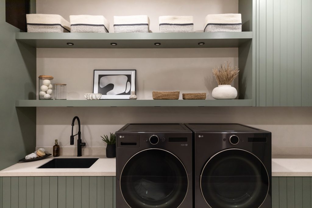 Laundry room with two front-load washers, open shelves holding towels and jars, and a sink with a faucet on a light countertop — Living with Lolo.