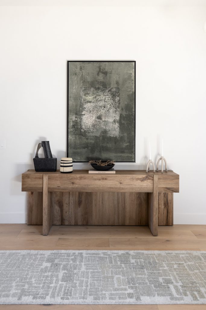 A wooden console table with decorative objects and two candlesticks stands beneath an abstract painting on a white wall—Living with Lolo.